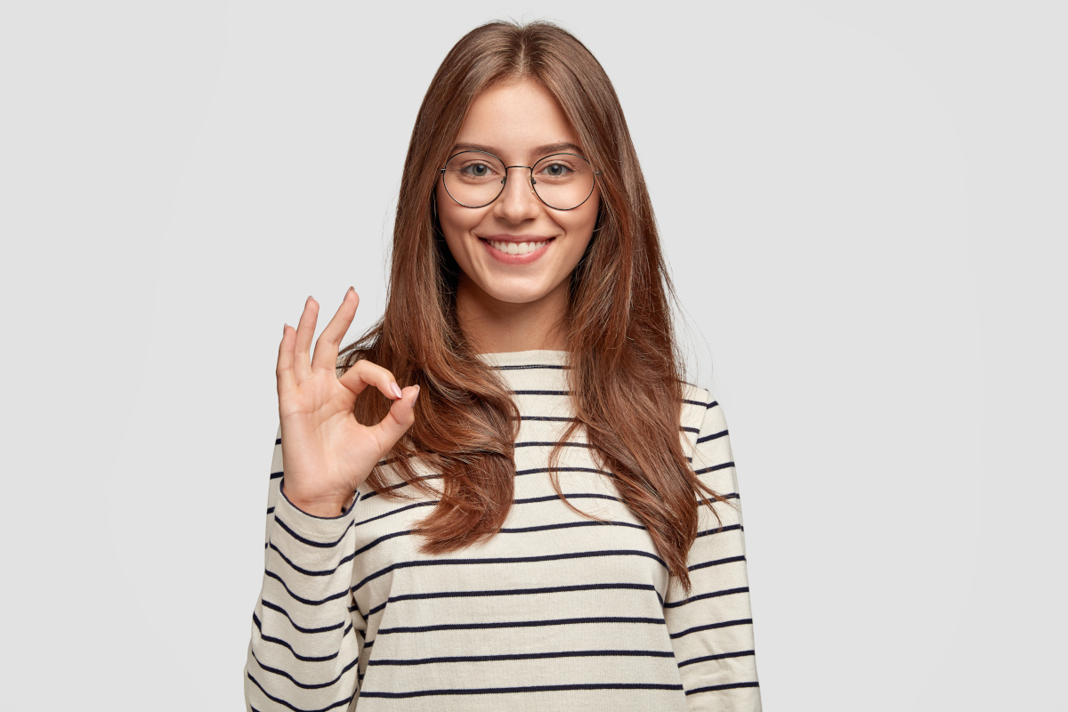 Dont worry, everything is good. Cheerful grinning young female in striped jacket, shows okay gesture, likes somebodys idea, poses against white studio wall. People and body language concept.
